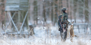 Ein Jäger steht vor einem Hochstand im verschneiten Wald und hält einen toten Fuchs