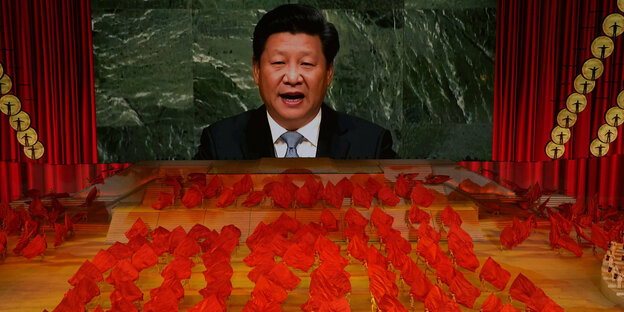 Xi Jinping auf einem Bildschirm , davor winzig TänzerInnen mit roten Fahnen
