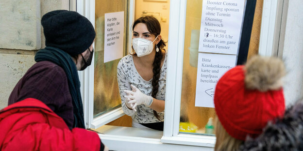 Eine Frau in weißer Kleidung und mit Maske lehnt sich aus einem Fenster im Erdgeschoss und spricht offenbar mit zwei Patienten.