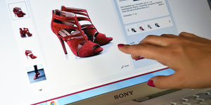 Eine Damenhand zeigt auf einen Computerbildschirm. Auf dem sieht man ein Paar rote, hochhackige Schuhe, die zum Verkauf angeboten werden.