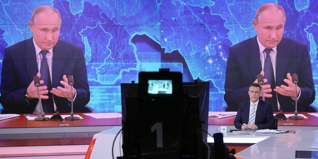 In einem Fernsehstudio sieht man einen Moderator, der vor einem großen Bildschirm sitzt, der Putin zeigt.