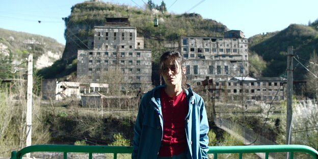 Eine Frau mit Jeansjacke und rotem Kapuzenpullover steht an einem Geländer und schaut erschöpft Richtung Boden, im Hintergrund sind alte Gebäude und die Taue der Seilbahnstrecke zu sehen