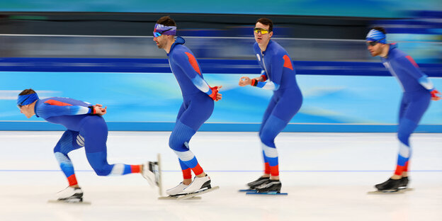 4 blau-weiß-rot-gekleidete Eisschnellläufer in einer großen Eissporthalle