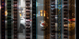 Filmstreifen laufen eine Glasscheibe herunter, in der Scheibe spiegeln sich der Bürgersteig und auf der Straße geparkte Autos