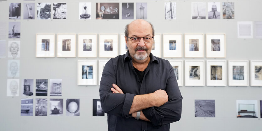 Ein Mann mit Brille und verschränkten Armen steht vor einer Wand, an der Kunstwerke hängen