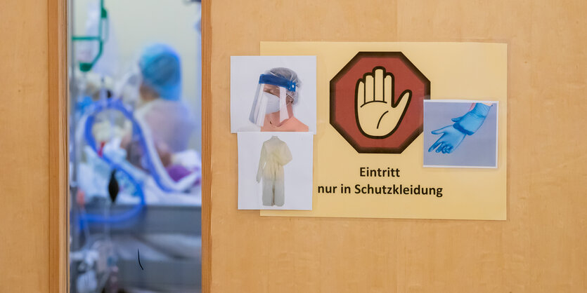 HInter einer etwas geöffneten Tür mit Corona-Hygienehinweisen ist ein Bett und eine Pflegeperson zu sehen