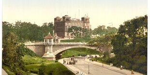 Die Deutsche Seewarte in einem nachcolorierten Foto. Das Backsteingebäude mit vier Türmen an den Ecken steht etwas erhöht, im Vordergrund eine Brücke und Fußgänger in einem Park.