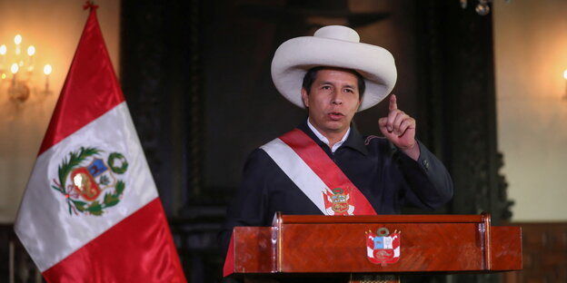 Perus Präsident Pedro Castillo mit großem weißen Hut und erhobenem Zeigefinger am Rednerpult neben der peruanischen Flagge