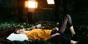 Eine Frau in einem gelben Pullover liegt auf einem Waldboden in Efeu und schaut in die Kamera. Ihr Kopf liegt auf einem Kopfkissen, hinter ihr sind weitere Beine auf dem Boden zu sehen, so wie die stehenden Beine von zwei Menschen, die Baustrahler halten