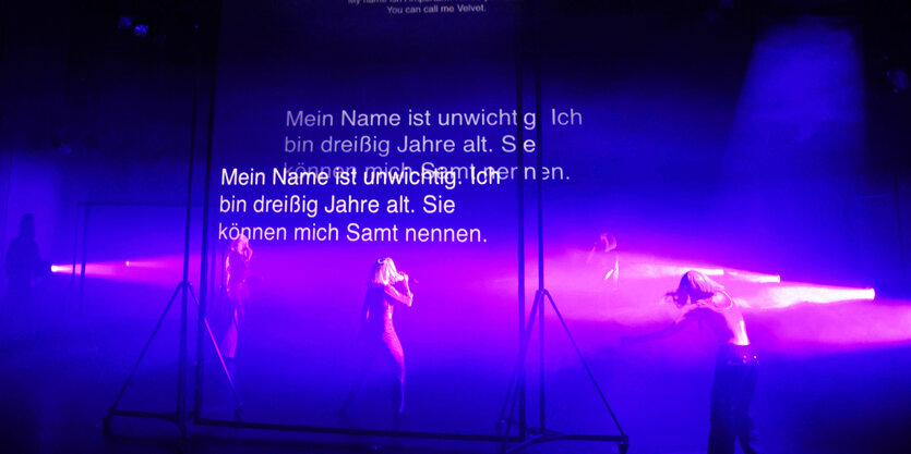 In blaues und lila Licht getaucht sind man Schauspieler und Schauspielerinnen auf der Bühne, über sie hinweg läuft Schrift