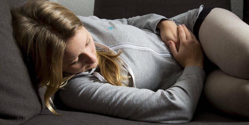 Eine junge frau liegt auf dem Sofa mit angezogenen Beinen und hält sich den Bauch vor Schmerzen fest