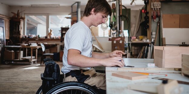 Ein junger Mann sitzt in einem Rollstuhl und arbeitet in einer Tischlerwerkstatt