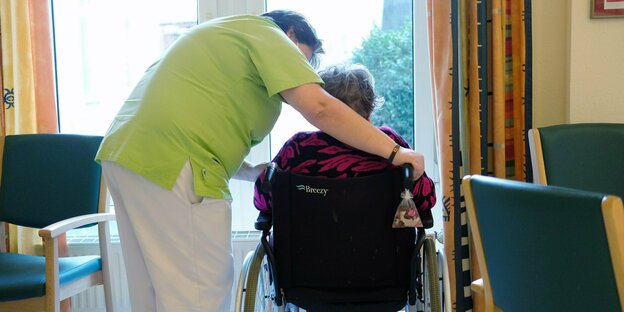 Eine Pflegerin bückt sich über einen Menschen im Rollstuhl - beide sind nur von hinten zu sehen