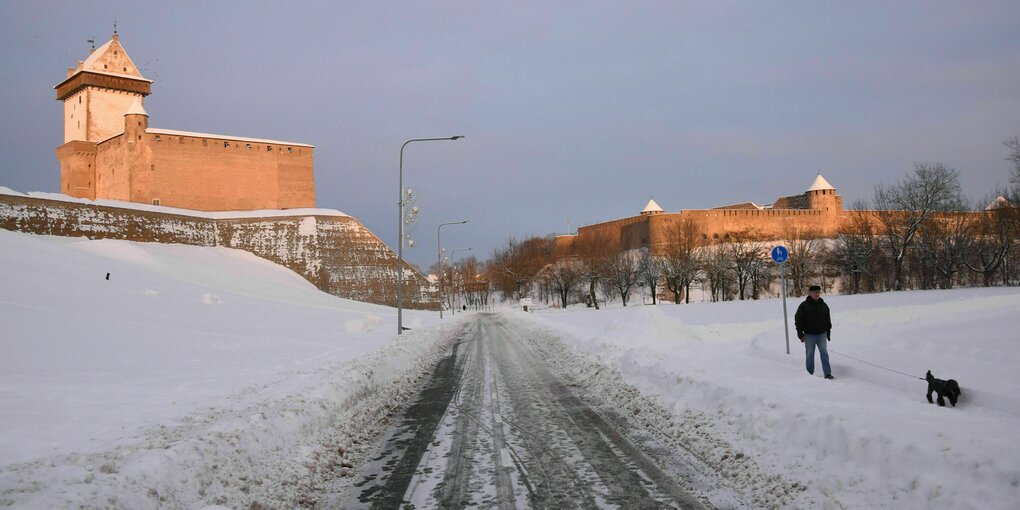 Ein Mann mit Hündchen im Schnee, dahinter ein Gebäude in der Sonne