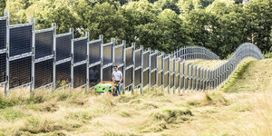 Senkrecht stehende Solarmodule, ein Mann mäht davor das Gras mit einer Maschine