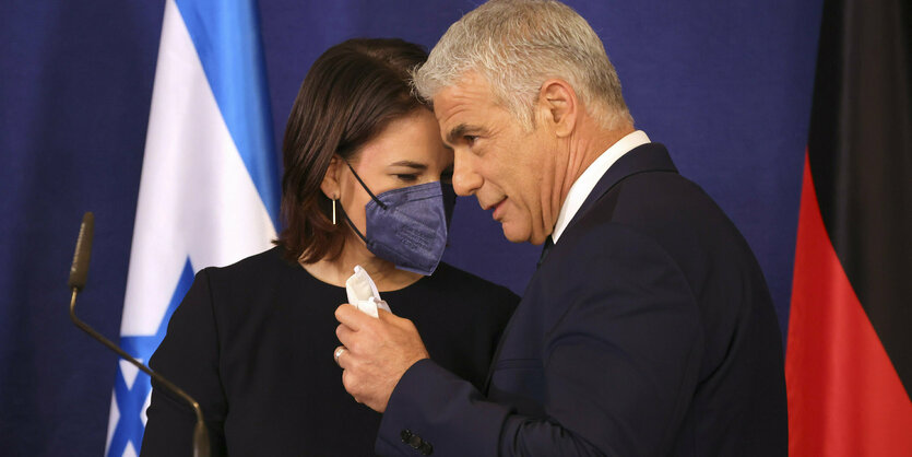 Annalena Baerbock mit Maske und Yair Lapid (setzt die Maske gerade auf) auf einer Pressekonferenz. Sie neigen sich einander zu, als sagten sie etwas zueinander, dabei berühren sich die Köpfe fast