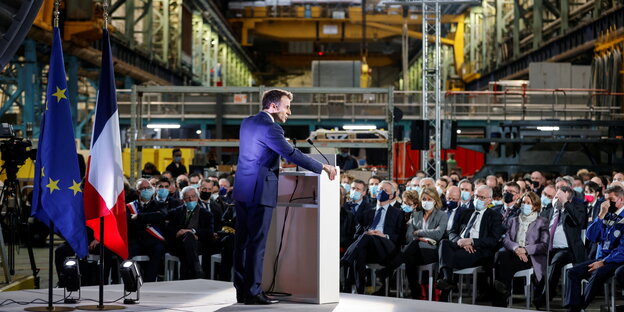 Präsident Emmanuel Macron spricht vor Menschen in einer Werkhalle.