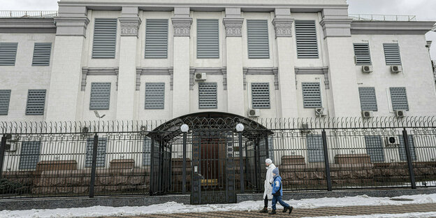 Ein großes weißes Gebäude mit geschlossenene Rolläden, ein Zaun und zwei Menschen, die vorbeilaufen