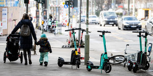 Städtische Szene: Mitten auf einem Bürgersteig stehen E-Scooter, eine Frau mit Kinderwagen und Kind an der Hand geht vorbei, im Hintergrund sieht man die Straße und eine Baustelle.