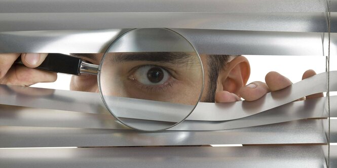 Person mit Vergrößerungsglas schaut durch Jalousie.