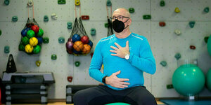 Ein Mann mit Mundschutzmaske sitzt auf einem Gymnastikball und fasst sich an Bauch und Brust
