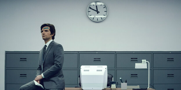 Szene aus der Serie: Mark (Adam Scott) sitzt im Anzug auf einem Schreibtisch, auf seiner Stirn prankt ein große blaues Pflaster