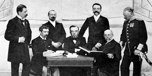 Baron Pierre de Coubertin und weitere IOC-Mitglieder haben sich um einen Tisch herum angeordnet