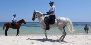 Zwei Sicherheitskräfte auf Pferden am Strand, im Hintergrund eine Touristin im Bikini..
