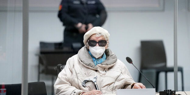 Irmgard Furchner sitzt im Rollstuhl, mit Mütze, OP-Maske und Sonnenbrille vor einer Justizbediensteten in Gerichtssaal