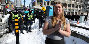 Auf einem schneebedeckten Bürgersteig in Ottawa: Im Vordergrund Frau im kurzärmeligen Shirt, die betet. Im Hintergrund uniformierte Polizisten