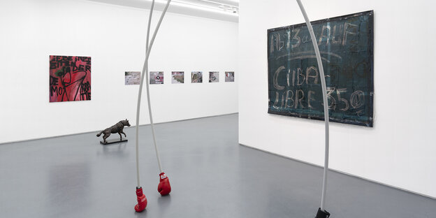 Ein paar rote Boxhandschuhe kommen als Teil einer Installation am Boden auf, dahinter ein dunkles Gemälde mit dem Schriftzug „Ab 13 Uhr auf / Cuba Libre: 3,50€“