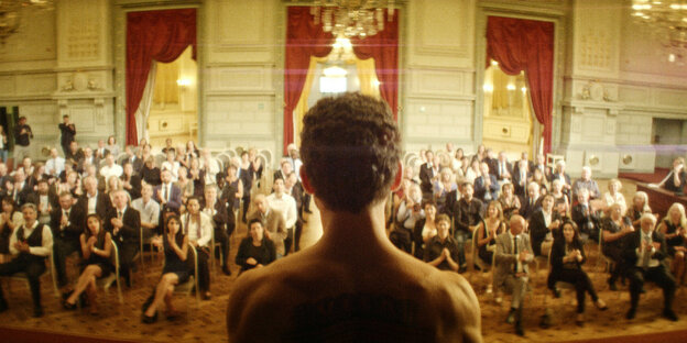 Junger Mann steht mit nacktem Oberkörper vor einem bürgerlichen Publikum