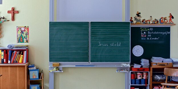 Ein Kreuz hängt neben der Tafel eines Klassenraums.