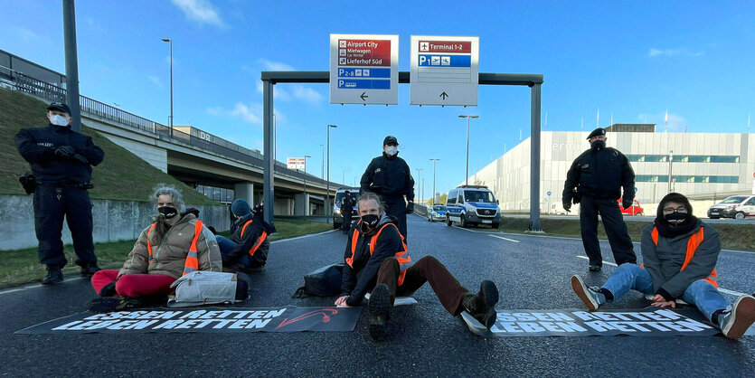 Junge Menschen sitzen mit Masken auf der Straße, bewacht von drei Polizisten