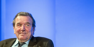Gerhard Schröder sitzt vor einer blau schimmernden Wand und lächelt