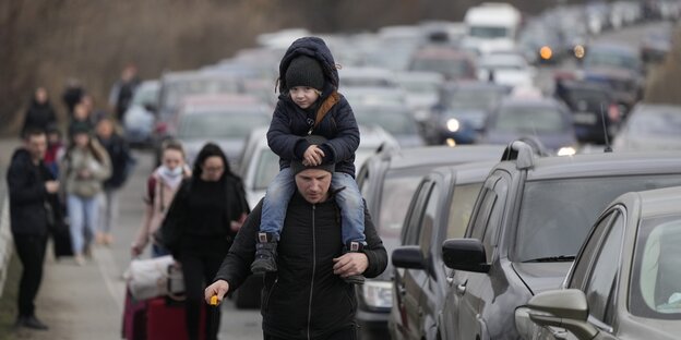Menschen fliehen, ein Vater trägt seinen Sohn auf der Schulter, viele Autos stehen im Stau