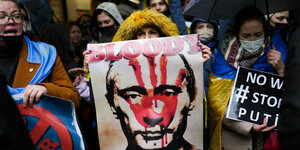 Eine Frau hält ein Plakat, auf dem Putin blutüberströmt zu sehen ist. Aufschrift "bloody killer". Andere Menschen rundum demonstrieren auch.