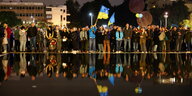 Menschen im Dunkeln demonstrieren und schwenken eine ukrainische Flagge, sie spiegeln sich in einem See
