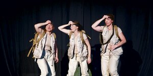 Drei als Soldaten verkleidete Schauspielerinnen stehen auf einer Bühne und fassen sich an den Kopf.
