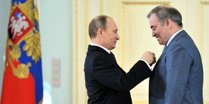 Eine russische Flagge, daneben Putin, der dem Komponisten Gergiyev einen Orden ansteckt, beide stehen sich gegenüber im Profil