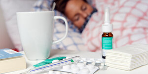 Eine Frau liegt krank im Bett. Vor ihr steht eine Tasse Tee, jede Menge Medikamente und Taschentücher.