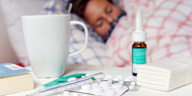 Eine Frau liegt krank im Bett. Vor ihr steht eine Tasse Tee, jede Menge Medikamente und Taschentücher.