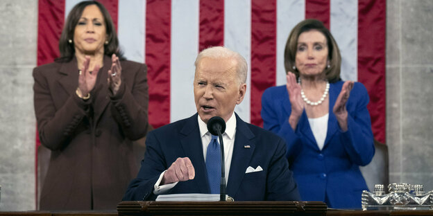 Joe Biden spricht am Podium, hinter ihm hängt die US-Flagge und zwei Frauen klatschen
