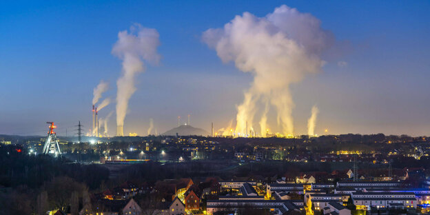Schornsteine eines kraftwerks stoßen Rauch aus, davor eine beleuchtete Stadt am Abend