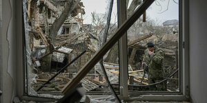 Blick aus einem zertörten Fenster auf einen zertrümmerten Hinterhof. Ein Soldat steht dort und wischt sich die Stirn ab, er wirkt extrem niedergeschlagen