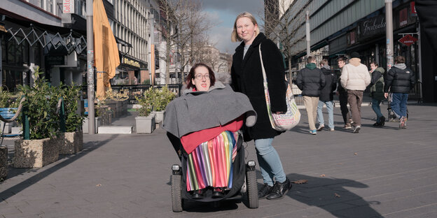 Victoria Michel ist im Rollstuhl, trägt einen bunten Rock. Daneben steht Alina Gerversmann. Sie sind draußen, neben ihnen sieht man Geschäfte und Fußgänger