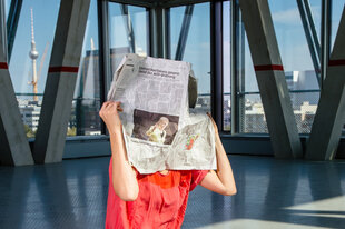 Im Haus der taz sitzt eine Frau hinter einer taz-Zeitung, ihr Gesicht ist von der Zeitung verdeckt. Im Hintergund erkennt man den Berliner Fernsehturm.