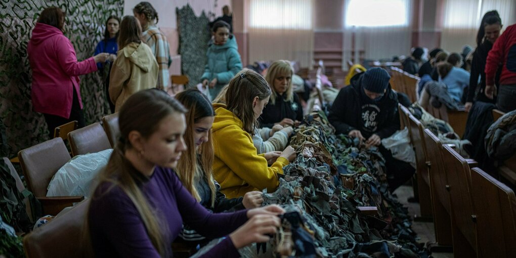 Frauen sitzen in einem Saal auf Klappsitzen und knüpfen Tarnnetze