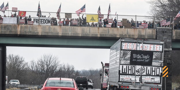 Ein mit US-Fahnen behängter und mit Sprüchen bemalter LKW fährt unter einer Brücke hindurch, auf der ebenfalls Menschen mit US-Fahnen winken.
