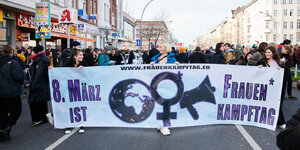 Die Frontansicht einer Demo; Demonstrierende halten ein weißes Transparent mit der Aufschrift: 8. März ist Frauenkampftag, daneben eine Weltkugel, ein Venusspiegel und ein Megafon